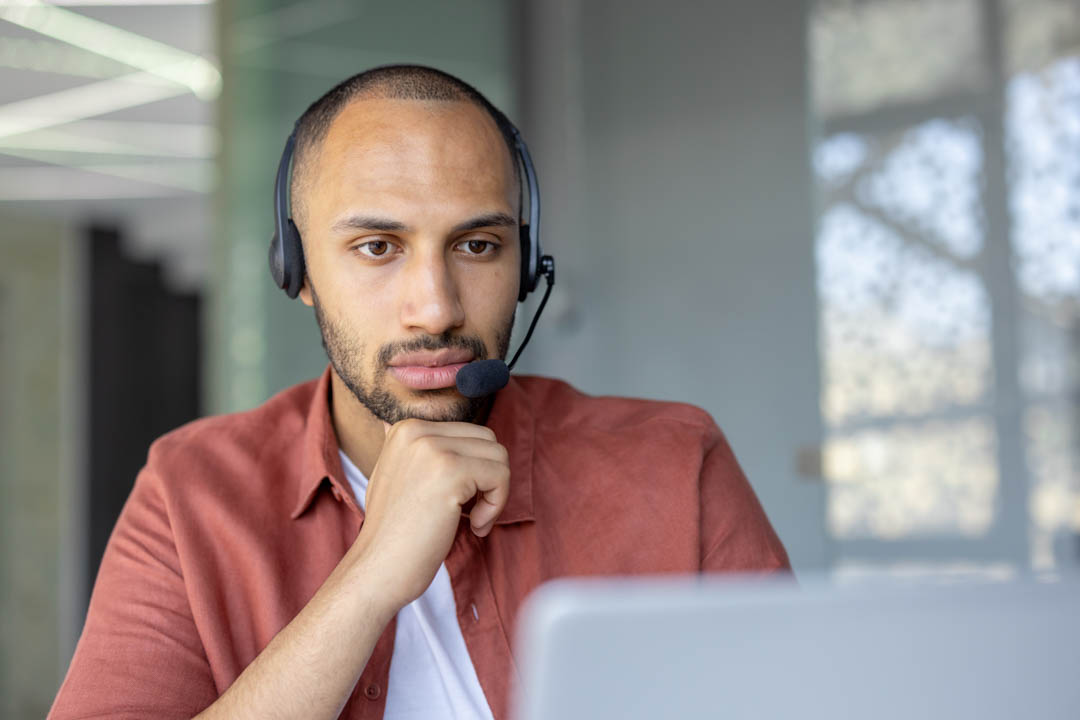 A thoughtful man wearing a headset, focused intently on his laptop screen. He's likely engaged in a virtual meeting or customer service call.