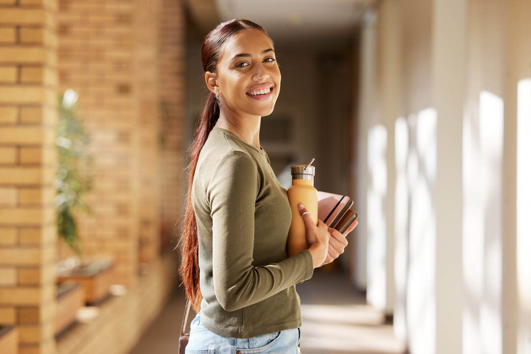 Woman portrait, university student and college books and water bottle while walking at campus with a smile. Gen z female happy about education, learning and future after studying at school building.